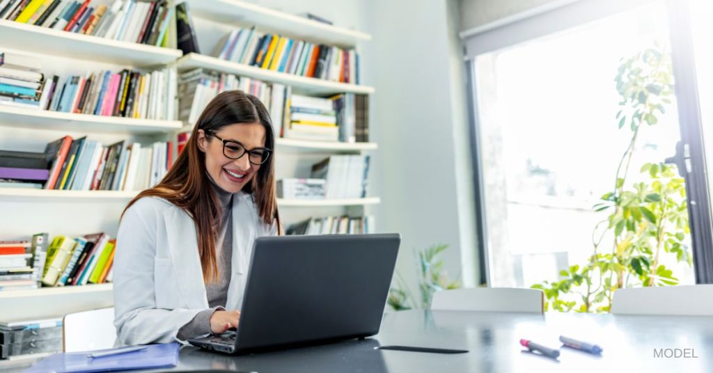 Woman in a white coat and glasses sitting in front of a laptop smiling. (MODEL)